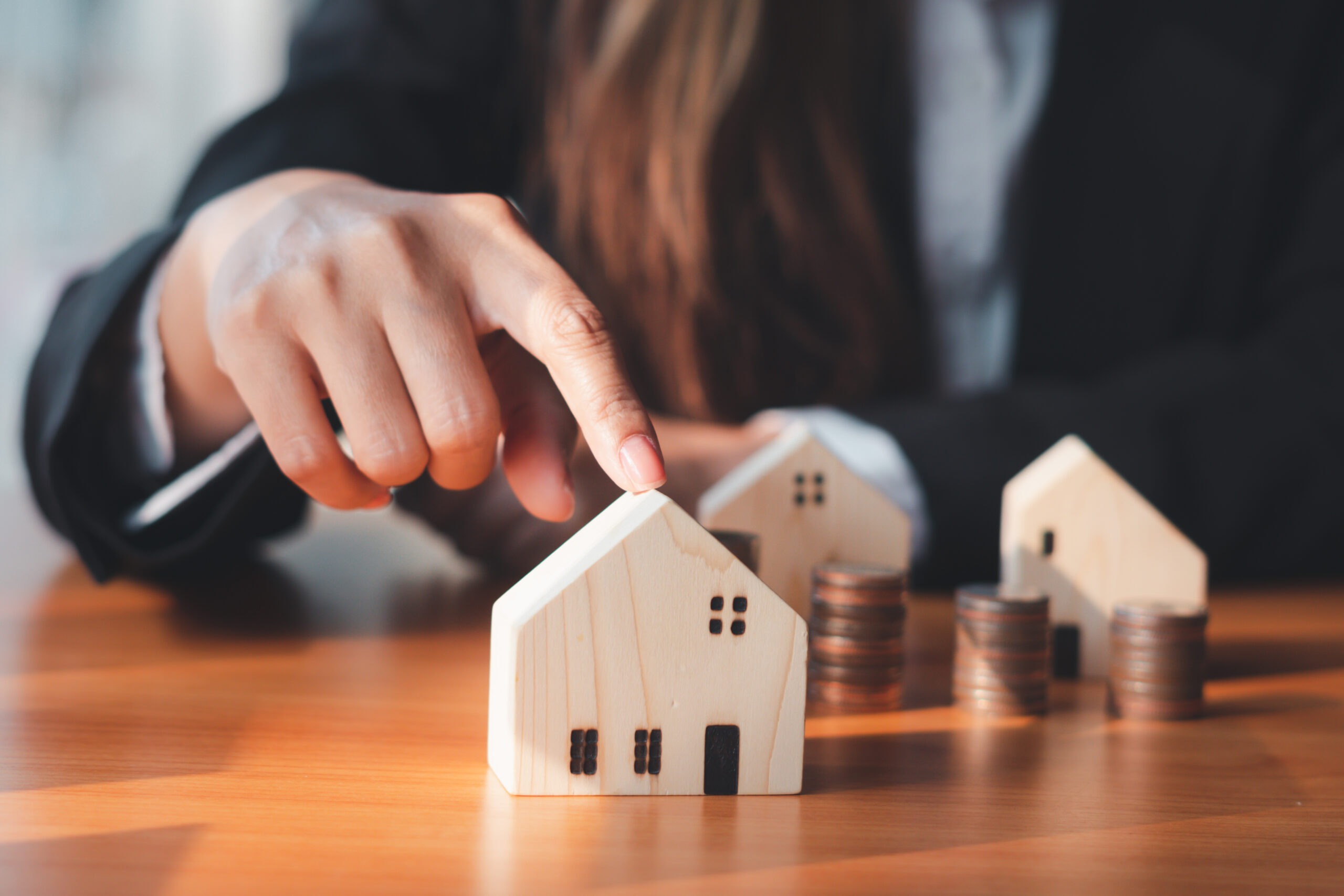 A hand points to a wooden house model, accompanied by stacks of coins